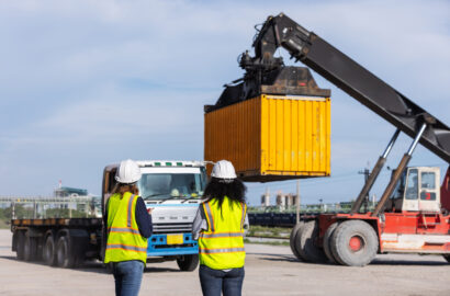 Two female workers in safety vests and hard hats oversee container loading operations at a shipping yard. A crane lifts a yellow container onto a truck, highlighting teamwork and global logistics.